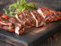 Rosticciana al forno morbida, succulenta e glassata, servita su tagliere scuro con pomodorini e rosmarino.