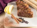 Fette di plumcake mela e cannella su piatto bianco, con mele, cannella, gocce di cioccolato e cacao in polvere.