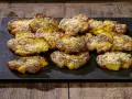 Patate schiacciate al forno, dorate e croccanti, cosparse di formaggio grattugiato e spezie, servite su una pietra scura.