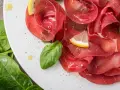 Fette sottili di bresaola di manzo su un piatto bianco, guarnite con spinaci freschi, spicchi di limone e un filo d'olio.
