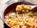 Un cucchiaio solleva una porzione fumante di pasta e patate veloce, filante di formaggio fuso, in una ciotola di terracotta.