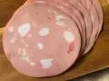Fette di mortadella su tagliere di legno. Scopri come si fa la mortadella, un salume rosa con cubetti di grasso bianco.