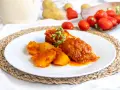 Piatto di cosciotto di tacchino in umido con patate, guarnito con erbe aromatiche. Sullo sfondo, pomodori e patate fresche.