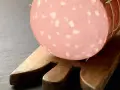 Una fetta di mortadella è cotta, con i suoi tipici cubetti bianchi di grasso, posata su un tagliere di legno.