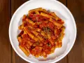 Un piatto di pasta alla pizzaiola, con rigatoni conditi con sugo di pomodoro, capperi e origano, servito in una ciotola bianca su un tavolo di legno scuro.