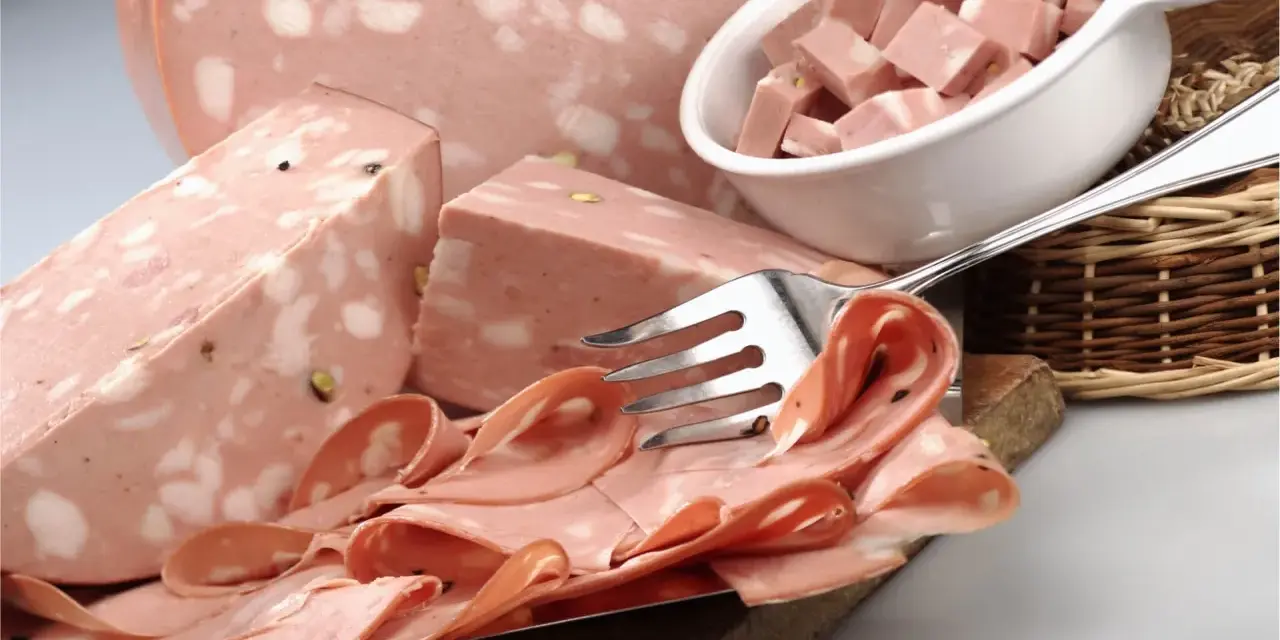 Fetta di mortadella con pistacchi e pepe, servita su un tagliere con fette arrotolate e cubetti in una ciotola.