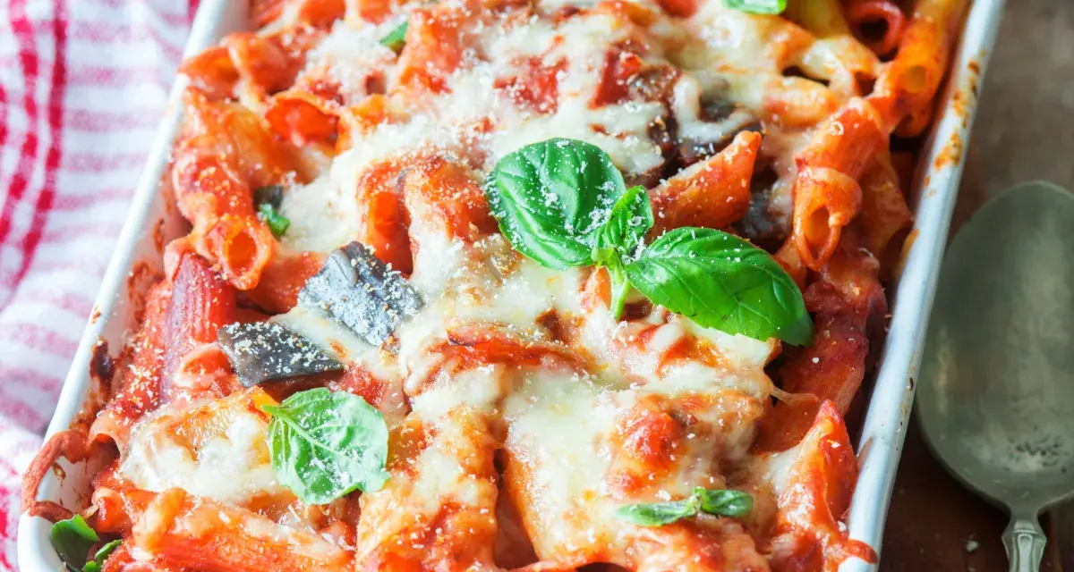 Piatto fumante di pasta al forno con melanzane, pomodoro e abbondante formaggio fuso, guarnito con foglie di basilico fresco.