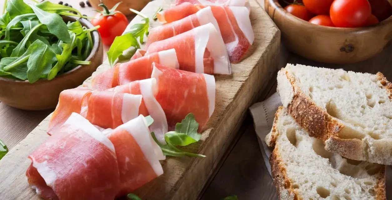 Fette di prosciutto crudo su tagliere con rucola, pomodorini e pane. Scopri come utilizzare il grasso del prosciutto crudo per insaporire i tuoi piatti.