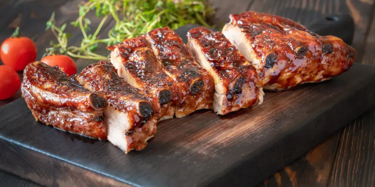 Rosticciana al forno morbida, succulenta e glassata, servita su tagliere scuro con pomodorini e rosmarino.