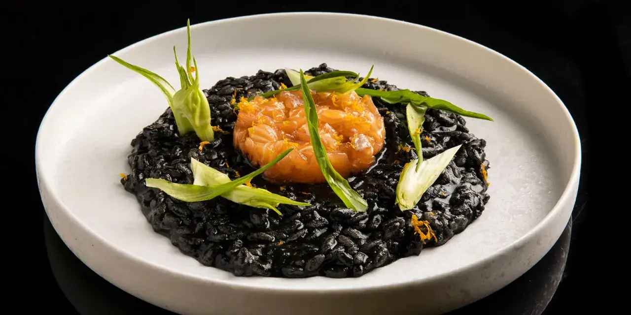 Un piatto di risotto al nero di seppia, guarnito con tartare di salmone e germogli verdi.