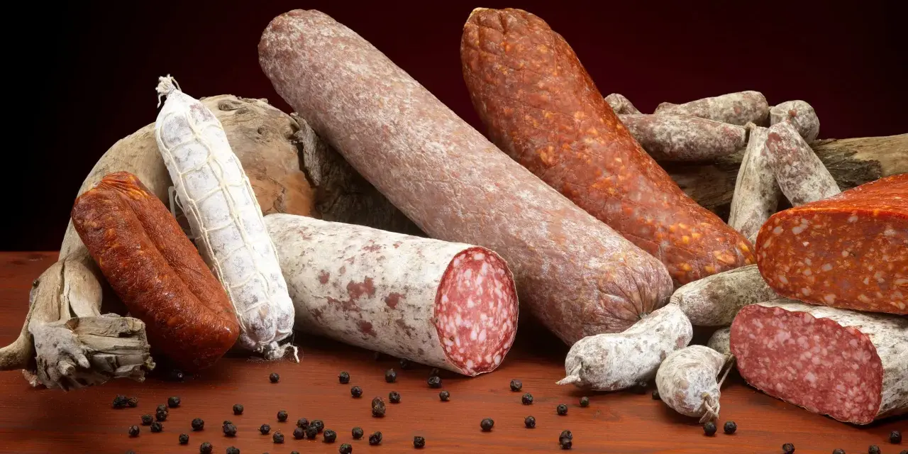 Un assortimento di salumi, tra cui spicca una grande fetta di carne insaccata, su un tavolo di legno con grani di pepe nero.