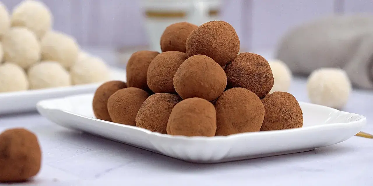 Montagna di tartufi al cioccolato senza panna, spolverati di cacao, su un piatto bianco. Sullo sfondo, altri dolcetti chiari.