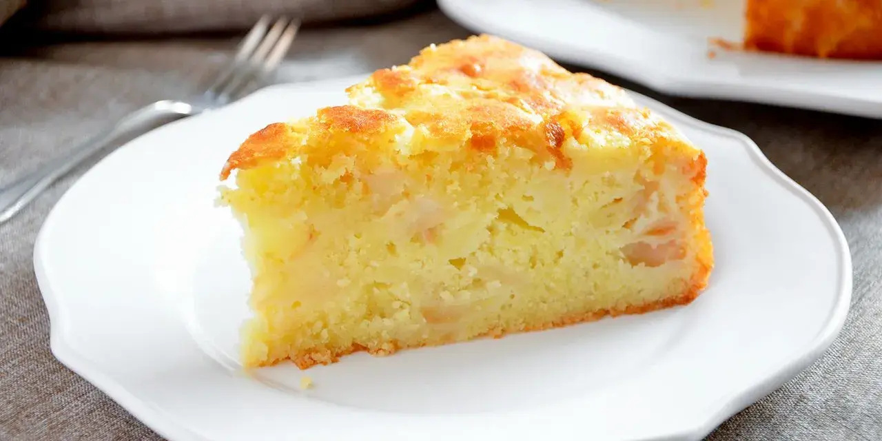 Una fetta di torta margherita con mele, soffice e dorata, servita su un piatto bianco.