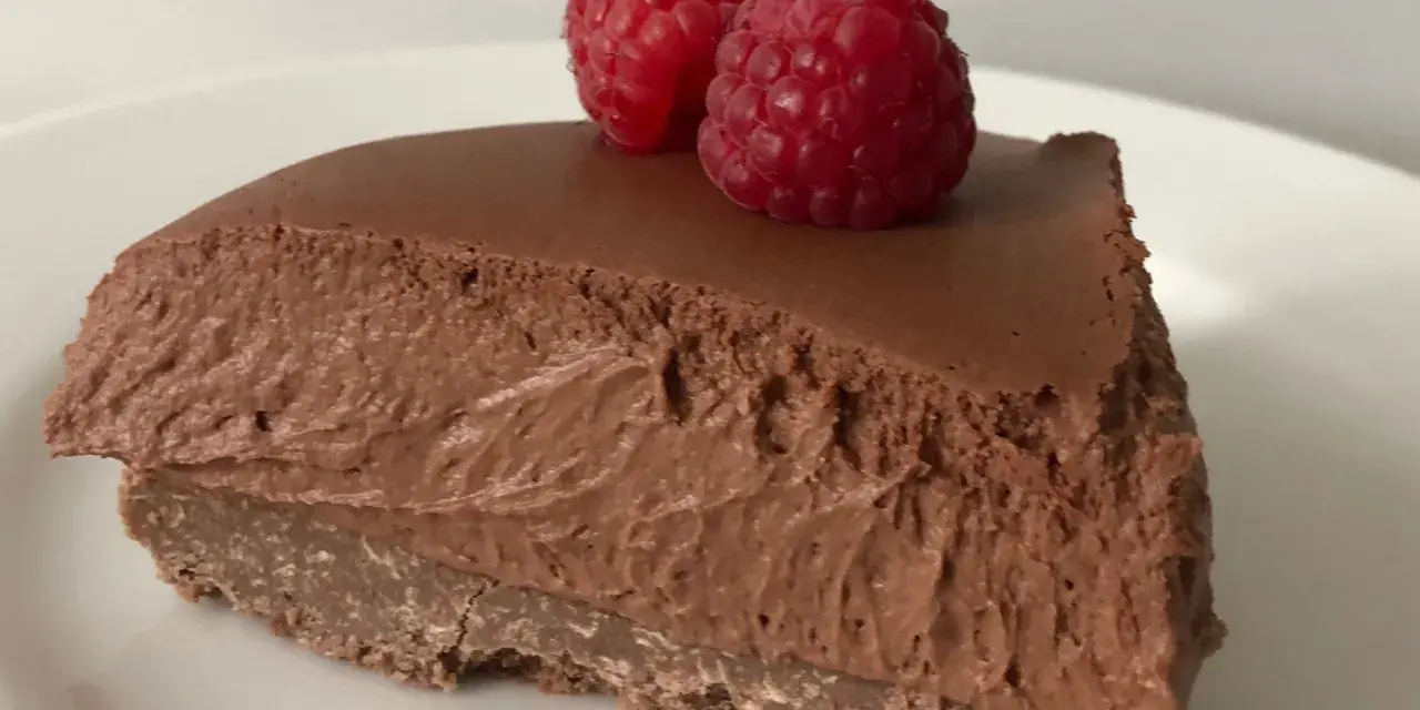 Una fetta di torta mousse al cioccolato, soffice e golosa, guarnita con due lamponi freschi su un piatto bianco.