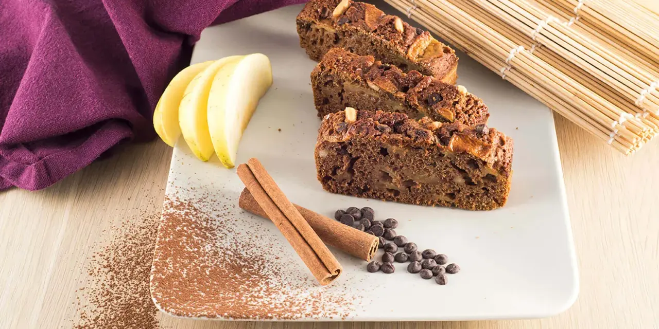 Fette di plumcake mela e cannella su piatto bianco, con mele, cannella, gocce di cioccolato e cacao in polvere.