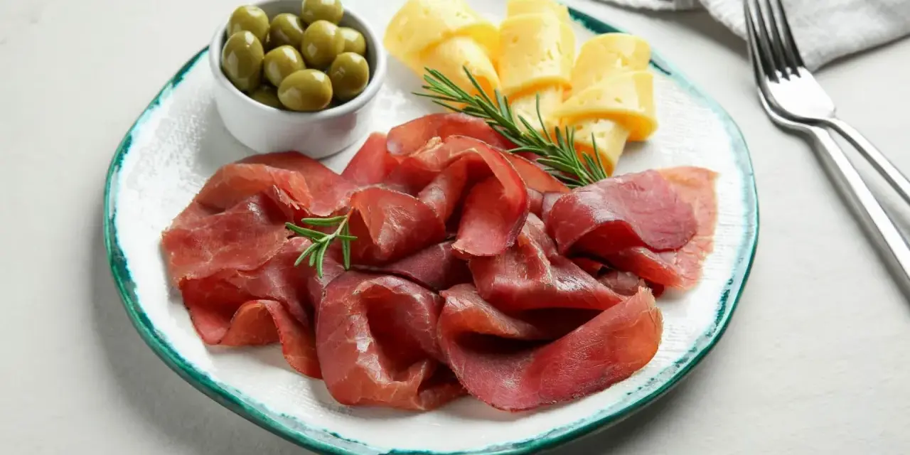 Bresaola con formaggio da abbinare, olive verdi e rosmarino su un piatto.