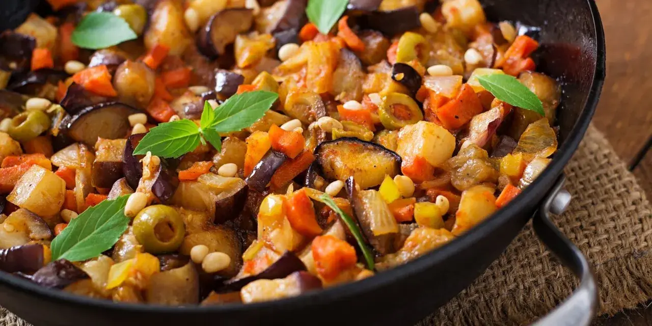 Caponata ricetta: melanzane, carote, olive e pinoli in padella, guarnita con basilico fresco. Un piatto colorato e saporito.