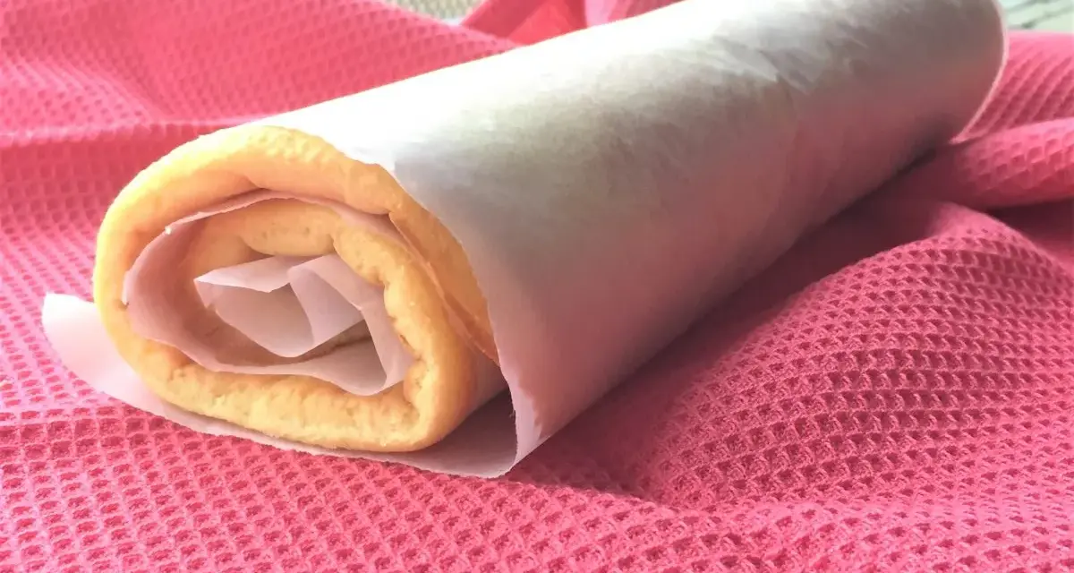 Rotolo di torta biscotto appena preparato, avvolto in carta forno, adagiato su un panno rosa.
