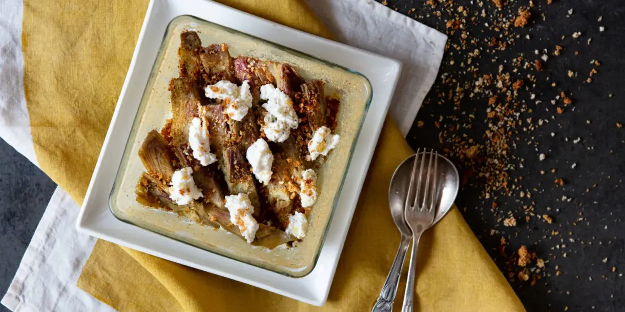Carciofi e ricotta al forno in una teglia bianca, guarniti con pangrattato croccante. Serviti su un tovagliolo giallo.