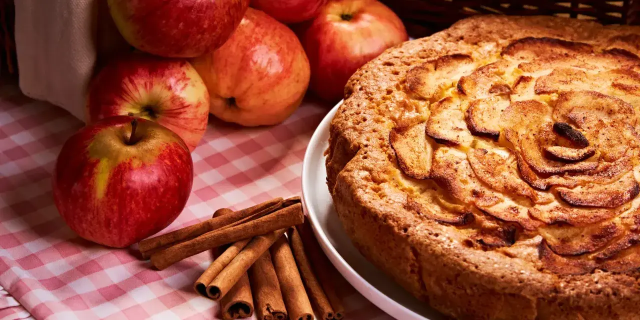 Torta mele, noci e cannella dorata, con mele fresche e bastoncini di cannella su una tovaglia a quadri rosa.