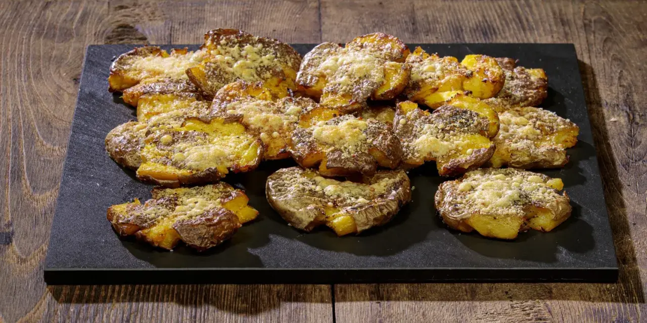 Patate schiacciate al forno, dorate e croccanti, cosparse di formaggio grattugiato e spezie, servite su una pietra scura.