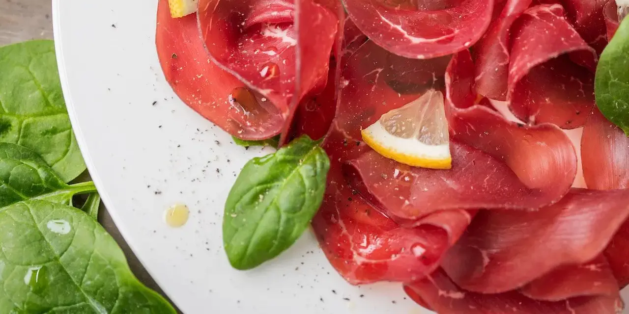 Fette sottili di bresaola di manzo su un piatto bianco, guarnite con spinaci freschi, spicchi di limone e un filo d'olio.