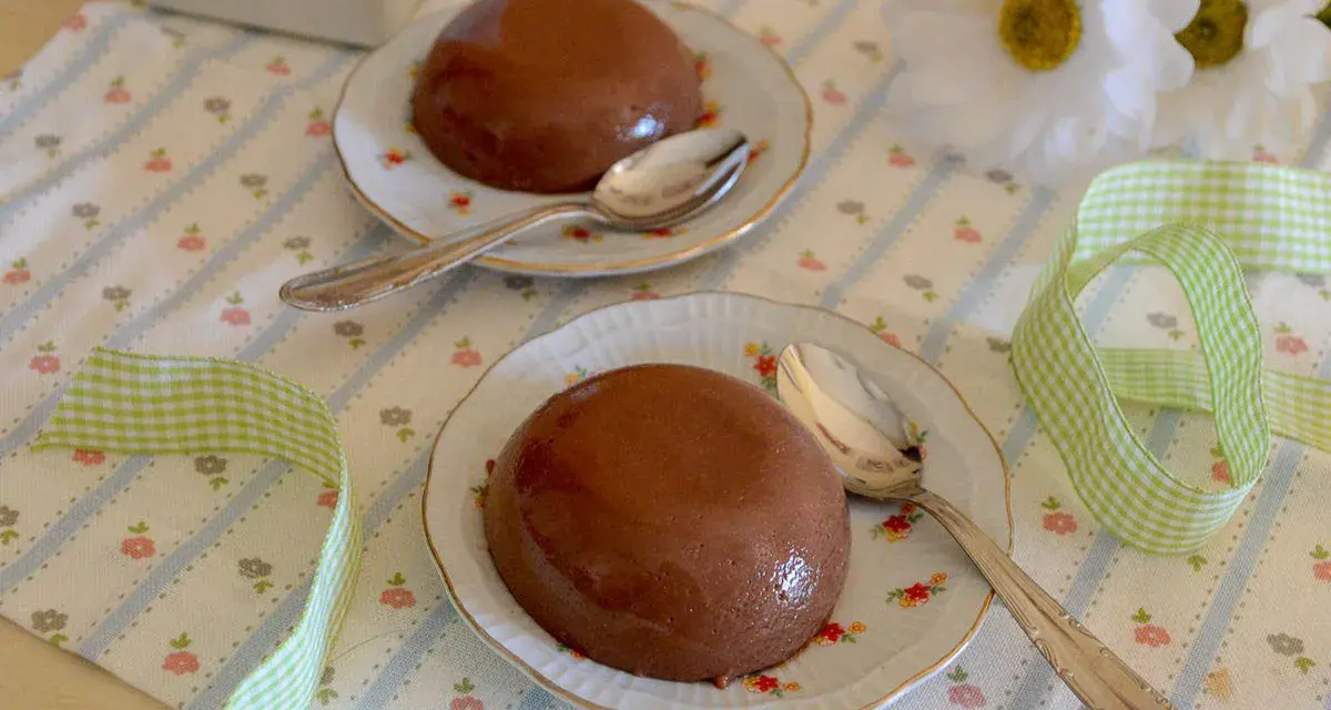 Due budini al cioccolato con amido di mais, serviti su piattini floreali con cucchiaini d'argento. Nastro a quadretti verdi completa la scena.