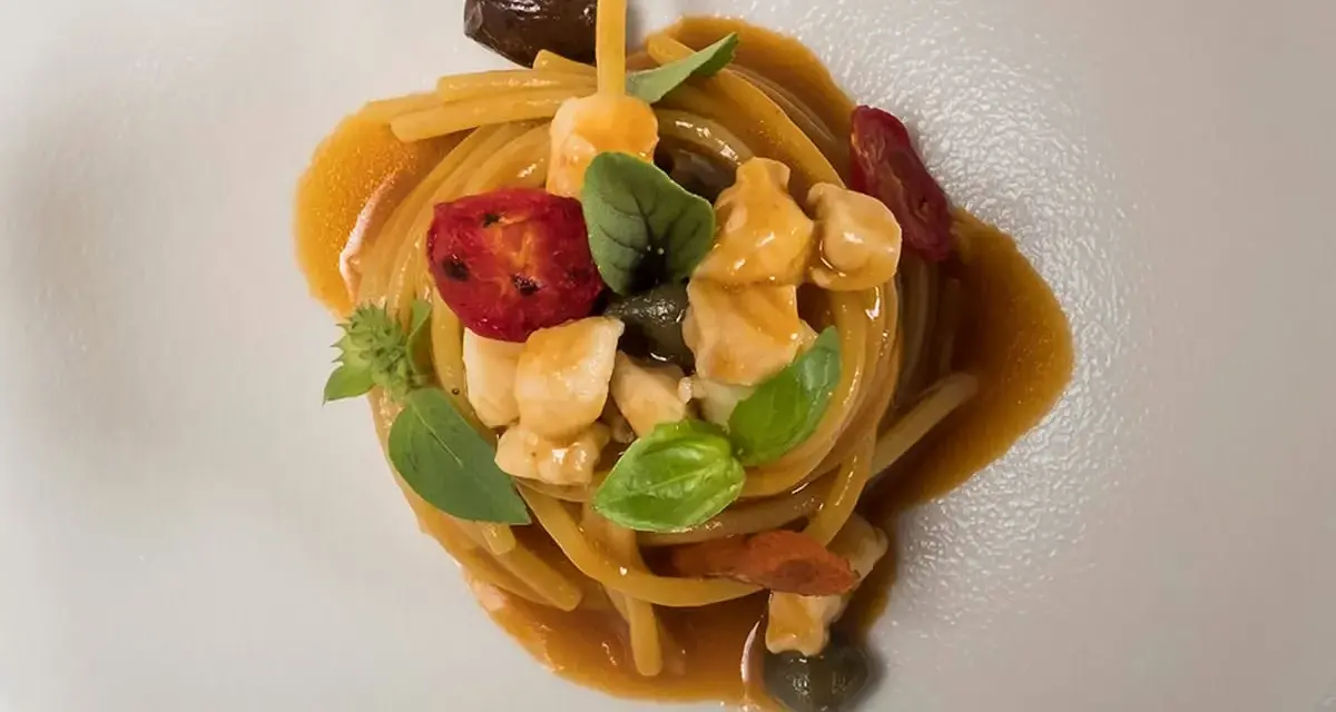 Un piatto di pasta con il pesce, condita con pomodorini, olive, capperi e basilico, servita su un piatto bianco.