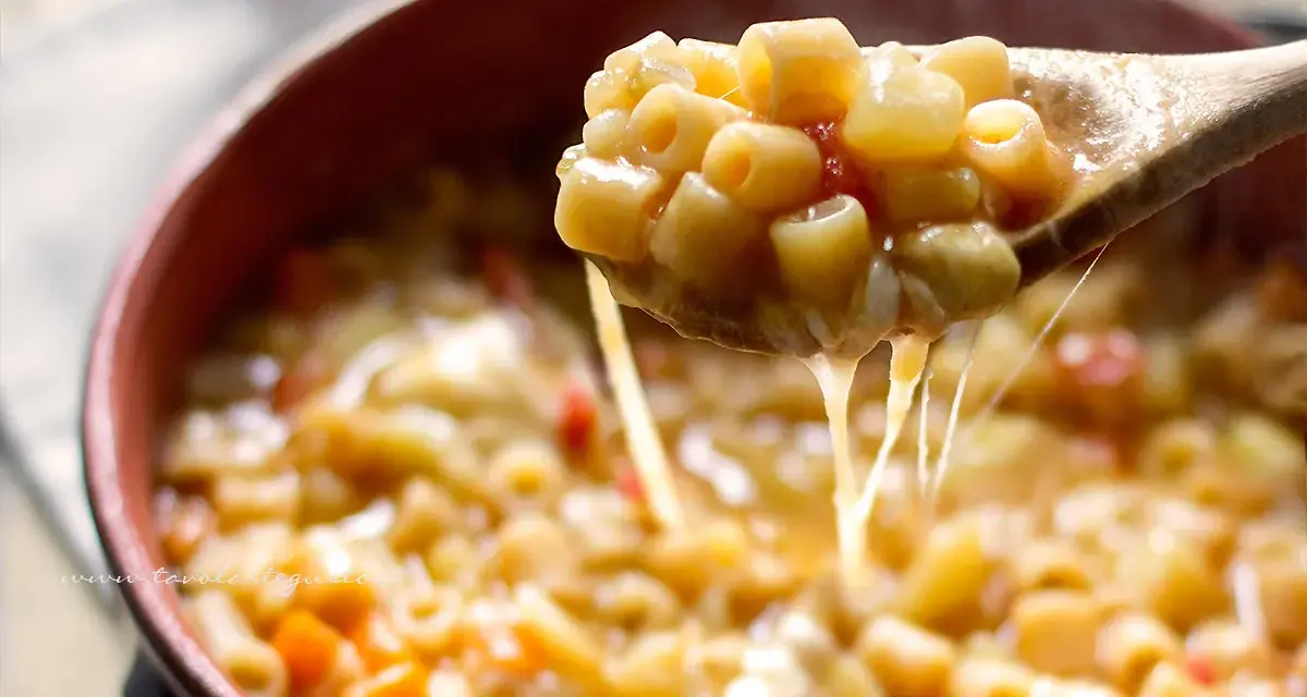 Un cucchiaio solleva una porzione fumante di pasta e patate veloce, filante di formaggio fuso, in una ciotola di terracotta.