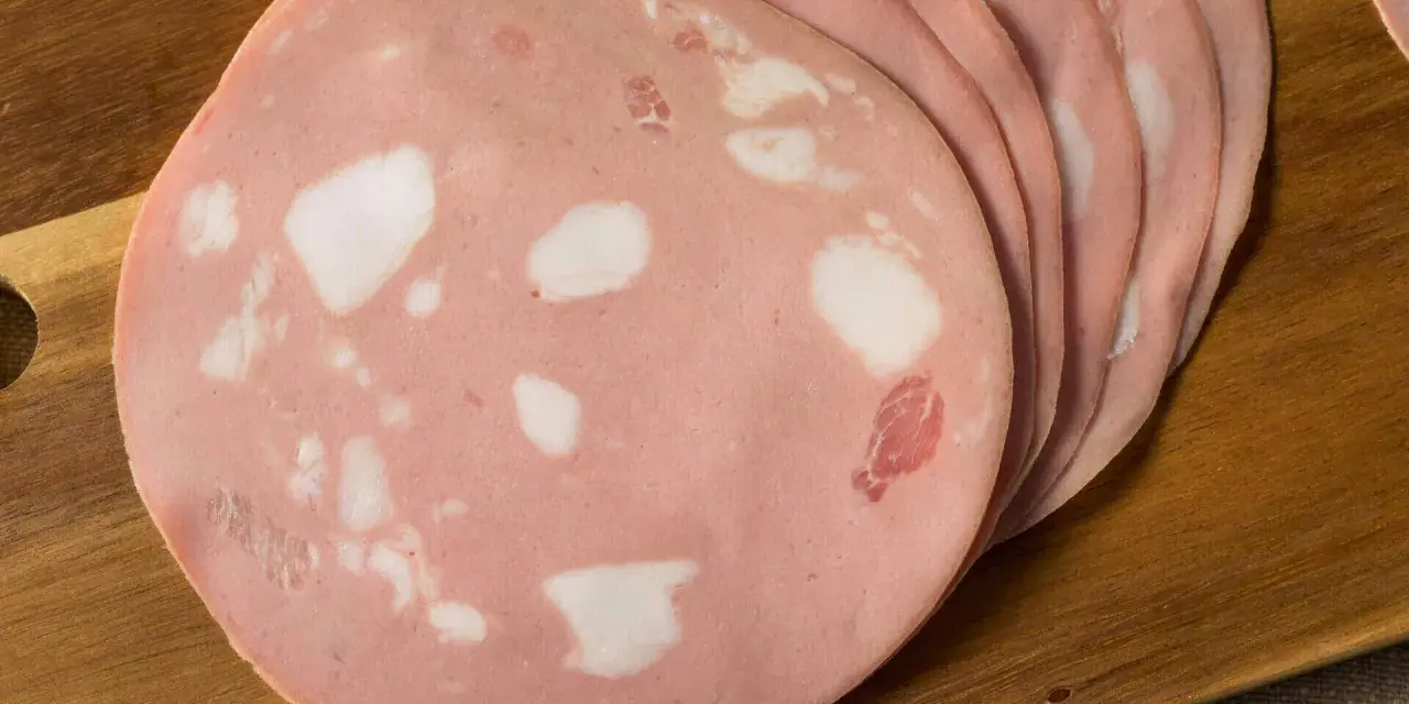 Fette di mortadella su tagliere di legno. Scopri come si fa la mortadella, un salume rosa con cubetti di grasso bianco.
