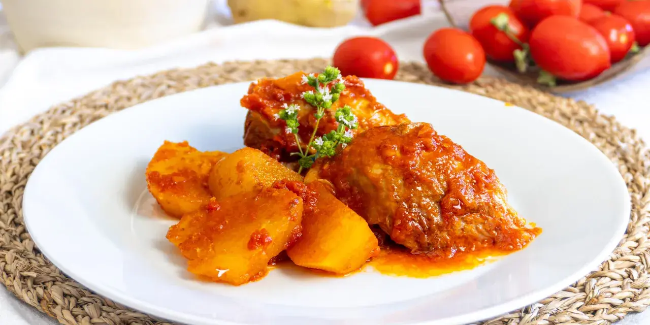 Piatto di cosciotto di tacchino in umido con patate, guarnito con erbe aromatiche. Sullo sfondo, pomodori e patate fresche.