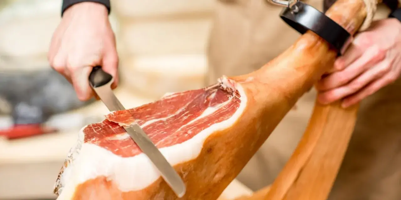 Mani esperte dimostrano come tagliare il prosciutto crudo con un coltello affilato, creando fette sottili e gustose.