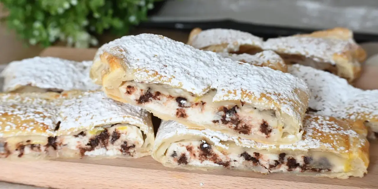 Rotolo di sfoglia con ricotta dolce e gocce di cioccolato, spolverato di zucchero a velo, su un tagliere di legno.