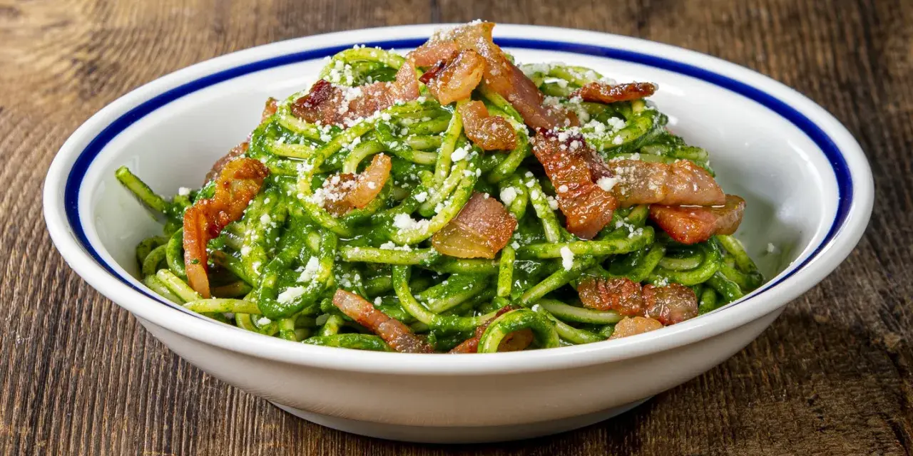 Un piatto invitante di pasta con spinaci, condita con croccante pancetta e parmigiano grattugiato, servita in una ciotola bianca con bordo blu.