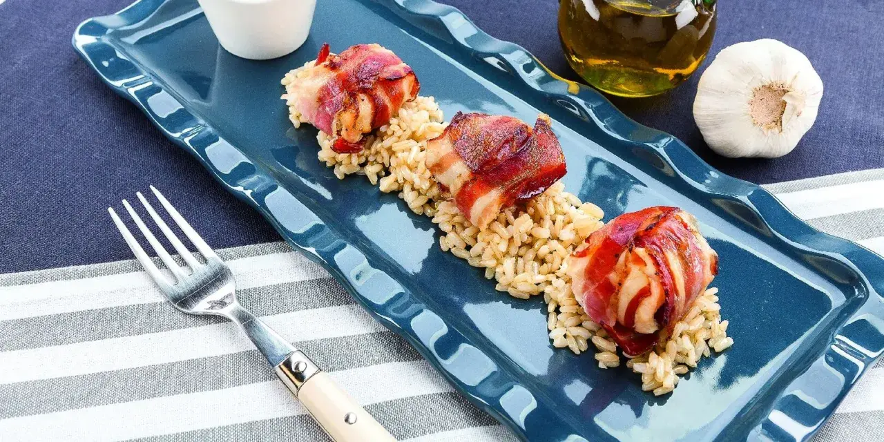 Tre bocconcini di pollo con pancetta avvolti e dorati, serviti su un letto di riso integrale.