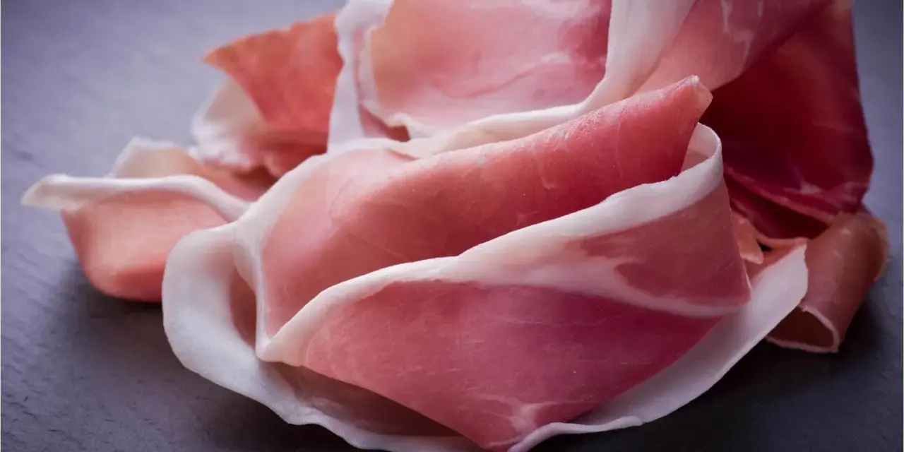 Fette di prosciutto crudo, un insaccato dal colore rosato e venature bianche, disposte su una superficie scura.
