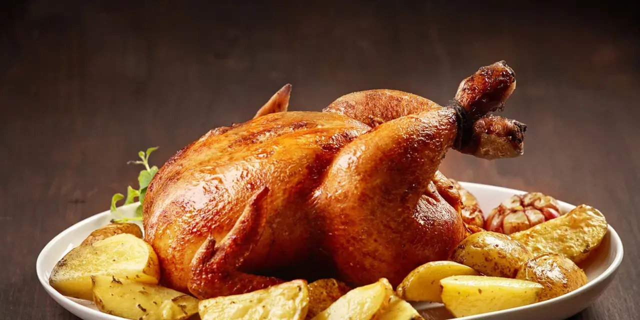 Un succulento pollo allo spiedo dorato, servito con patate arrosto e aglio su un piatto bianco.