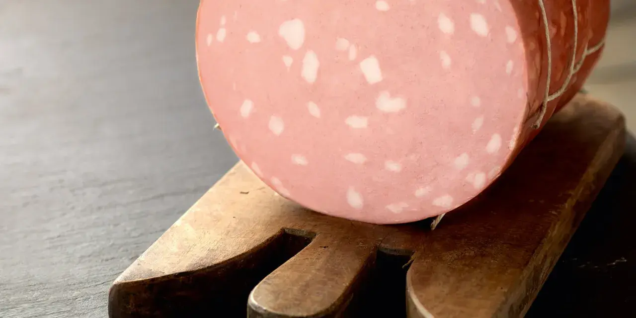 Una fetta di mortadella è cotta, con i suoi tipici cubetti bianchi di grasso, posata su un tagliere di legno.