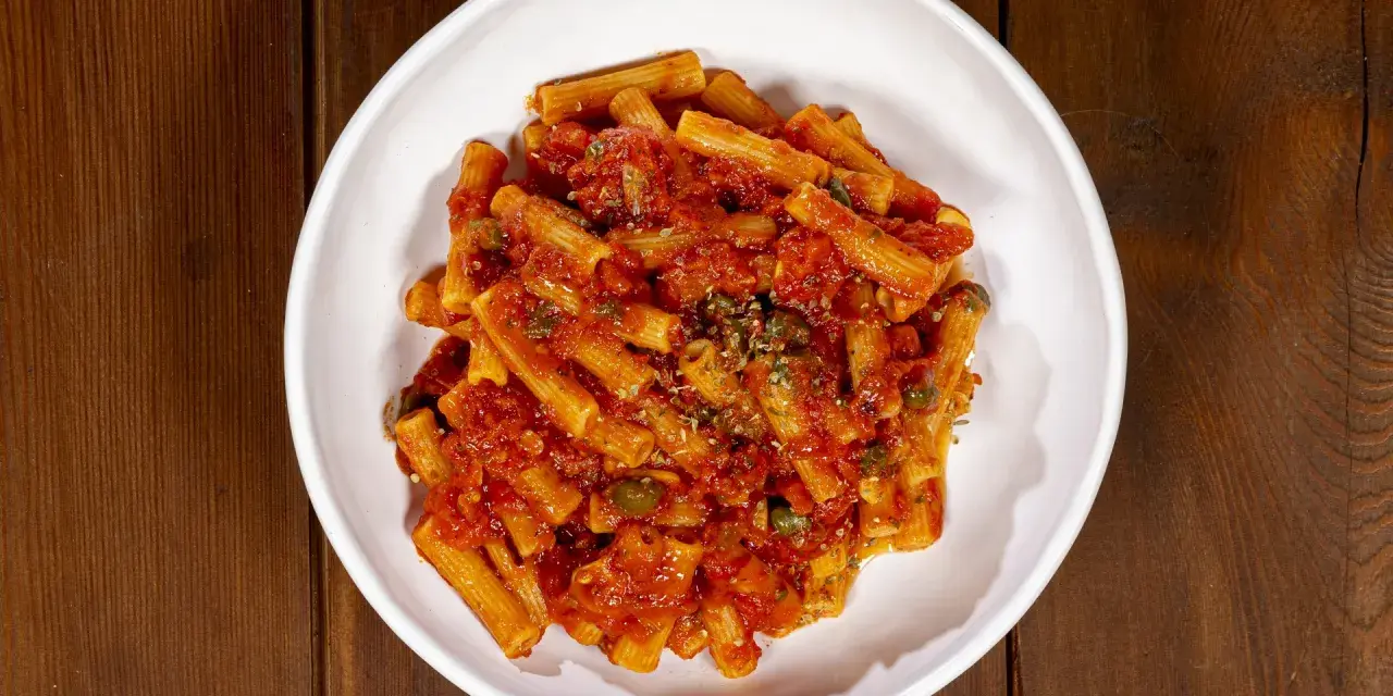 Un piatto di pasta alla pizzaiola, con rigatoni conditi con sugo di pomodoro, capperi e origano, servito in una ciotola bianca su un tavolo di legno scuro.