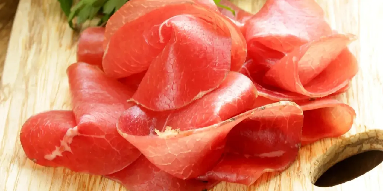 Fettine sottili di salume simile alla bresaola, rosse e lucide, disposte su un tagliere di legno con un ciuffo di prezzemolo.