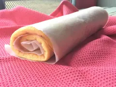 Rotolo di torta biscotto appena preparato, avvolto in carta forno, adagiato su un panno rosa.