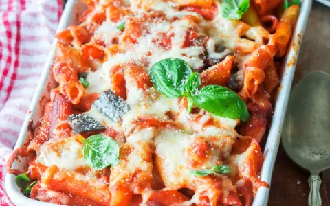 Piatto fumante di pasta al forno con melanzane, pomodoro e abbondante formaggio fuso, guarnito con foglie di basilico fresco.