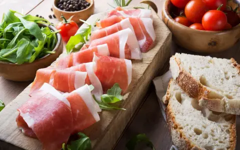 Fette di prosciutto crudo su tagliere con rucola, pomodorini e pane. Scopri come utilizzare il grasso del prosciutto crudo per insaporire i tuoi piatti.