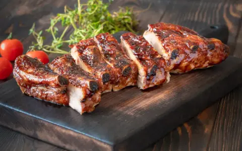 Rosticciana al forno morbida, succulenta e glassata, servita su tagliere scuro con pomodorini e rosmarino.