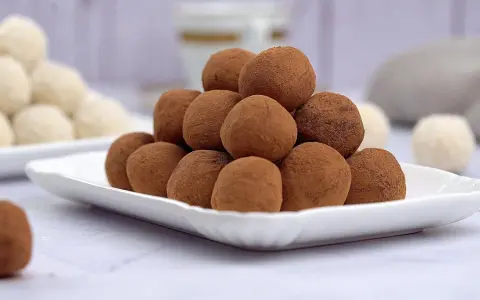 Montagna di tartufi al cioccolato senza panna, spolverati di cacao, su un piatto bianco. Sullo sfondo, altri dolcetti chiari.