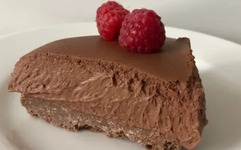 Una fetta di torta mousse al cioccolato, soffice e golosa, guarnita con due lamponi freschi su un piatto bianco.