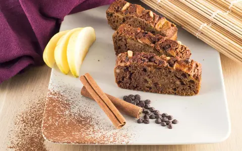 Fette di plumcake mela e cannella su piatto bianco, con mele, cannella, gocce di cioccolato e cacao in polvere.