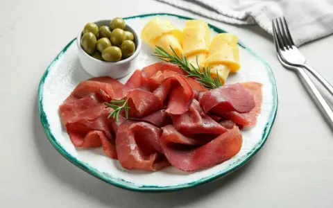 Bresaola con formaggio da abbinare, olive verdi e rosmarino su un piatto.