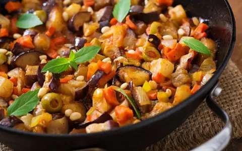 Caponata ricetta: melanzane, carote, olive e pinoli in padella, guarnita con basilico fresco. Un piatto colorato e saporito.