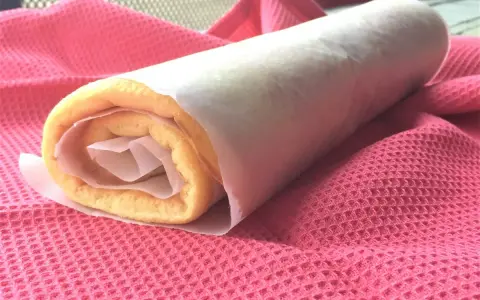 Rotolo di torta biscotto appena preparato, avvolto in carta forno, adagiato su un panno rosa.